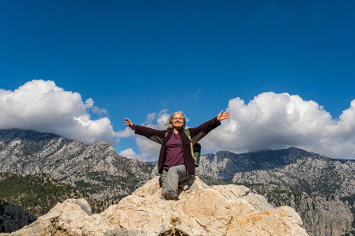 Old lady with backpack sitting on the rocks, laughing. Mountains and clouds in the background.