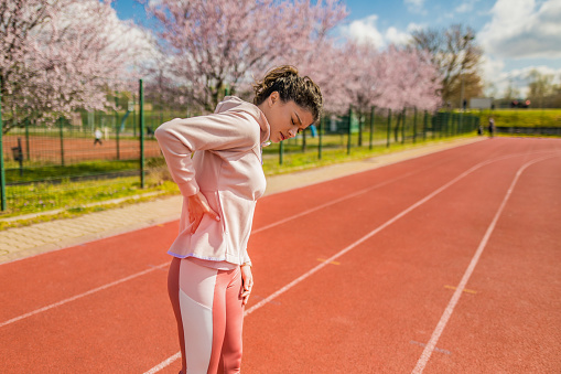 Unwell young female athlete having backache. She is exercising on sports track on early spring day.