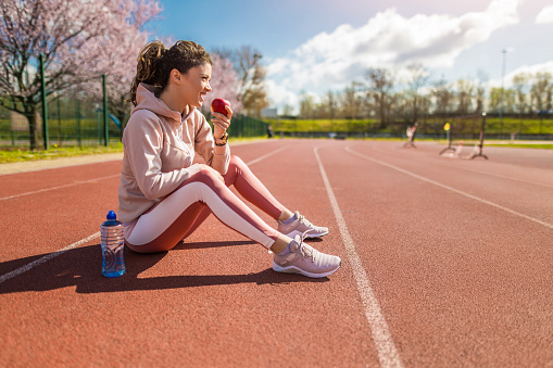 Young healthy female athlete eating an apple before sports training on running track. Beautiful vibrant colors on sunny early spring day and very high resolution.