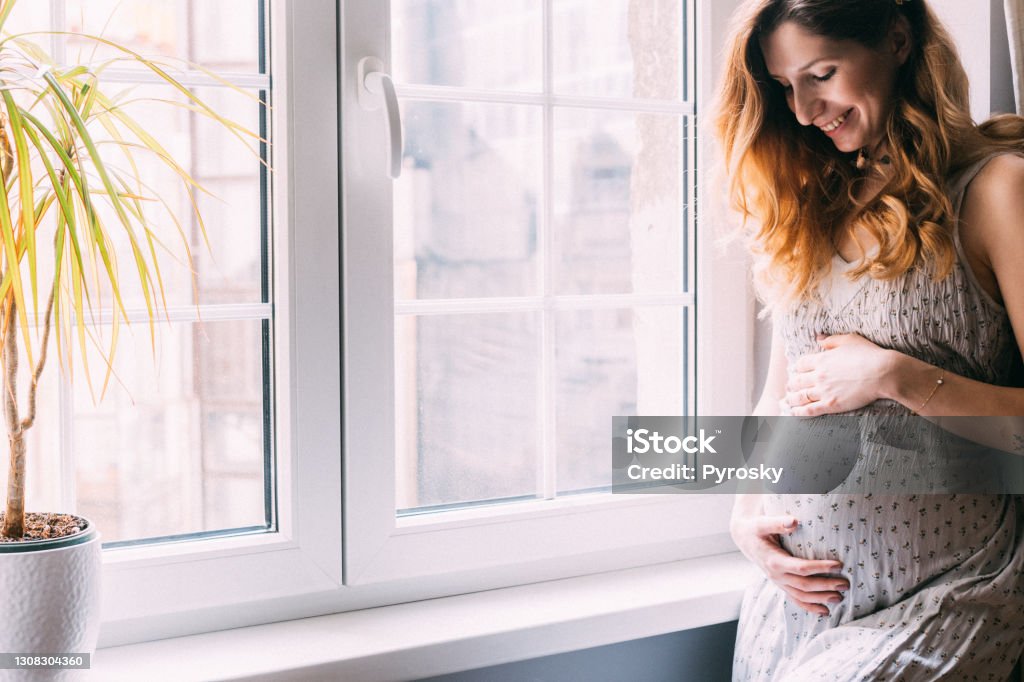 Portrait of a beautiful smilling pregnant woman Side view of a pregnant woman in a gray dress  holding her belly with a smile, standing by the window at home in a bright interior. Pregnant Stock Photo