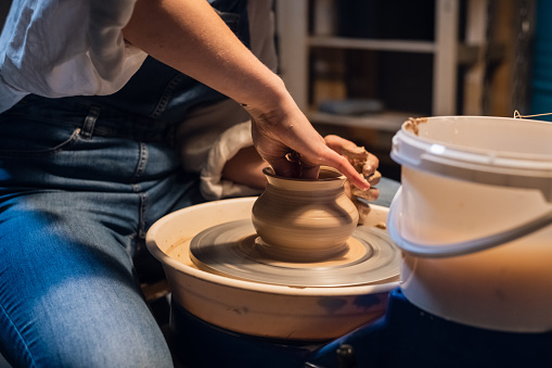 a young girl in a white blouse and denim overalls makes a pot on a potter's wheel with her hands