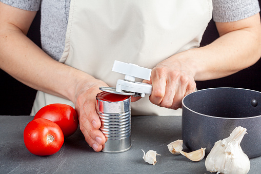 A woman is carefully opening a can of tomato paste on a kitchen counter using a white plastic can opener. She is preparing a meal for which she uses both fresh and pureed preserved tomatoes