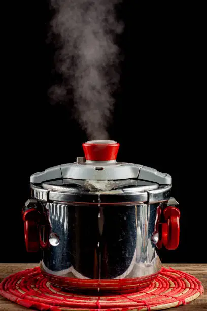 A steel pressure cooker is cooling on a fabric trivet on wooden table. The pressure valve is released and steam is coming out as the system gets depressurized. Hot steam leaving pot is seen.