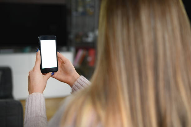 istock Back view close up of a woman hand holding and using smart phone with white blank empty screen sitting on a bed at home. 1308363821