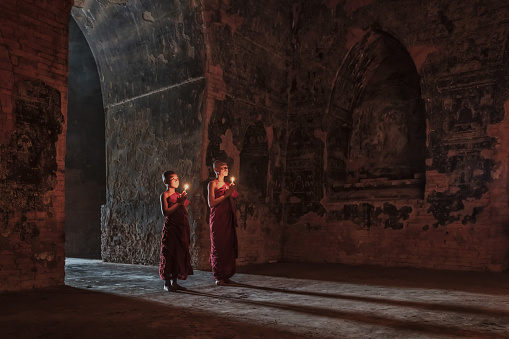Burmese Novice Monks standing side by side at the entrance inside dark buddhist temple. Holding candles, praying towards Buddha Statue. Moody Backlight from the temple entrance with long shadows. Tranquil Buddhist Worshipping Scene with Real People - Real Novice Monks. Old Bagan, Mandalay Region, Myanmar, Southeast Asia.
