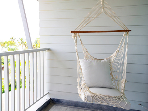 Empty white knitting rope swing hanging from ceiling on white wood background in room terrace in the resort. Vintage white handcraft hammock decoration on balcony.