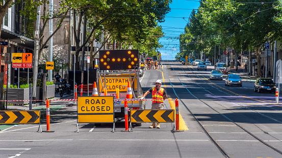 Melbourne, Victoria, Australia, January 31st, 2021: A male is wearing hi visibility clothing while manning a road closed station as workers perform roadworks in the background