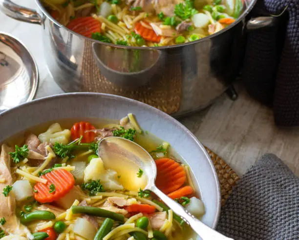 fresh and homemade cooked chicken soup with spring vegetables such as green pes, beans, carrots, caulifloer and carrots served in a bowl with spoon from above