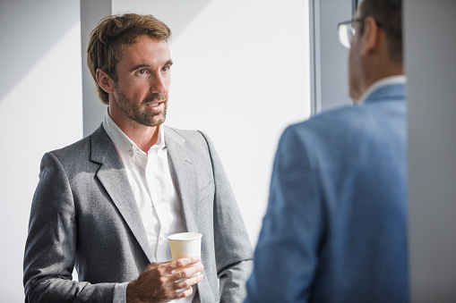 Young businessman standing by the big window and talking to senior colleague over coffee