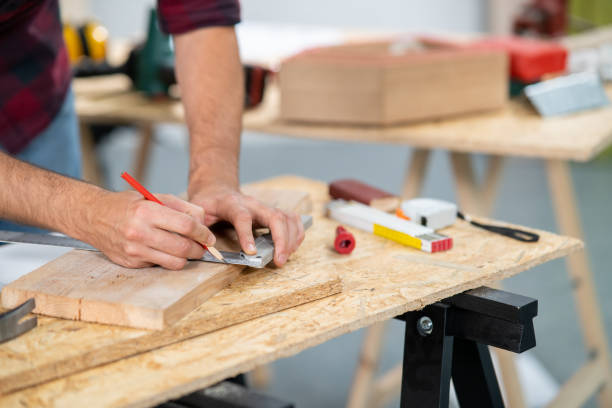 istock Close-up of a hardworking professional carpenter holding a angular ruler and pencil while measuring a board in a carpentry workshop. A bearded DIY enthusiast measures wood. There are a locksmith table and tools in the background 1308386791