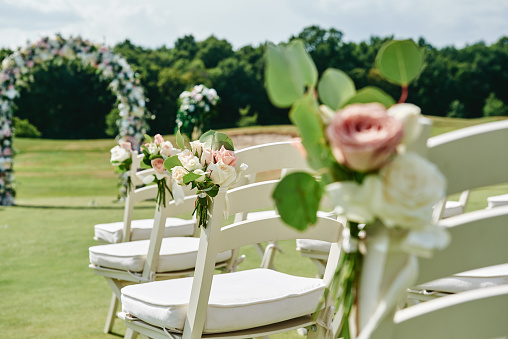 White wooden chairs with rose flowers on each side of archway outdoors, copy space. Empty chairs for guests prepared for wedding ceremony on golf course