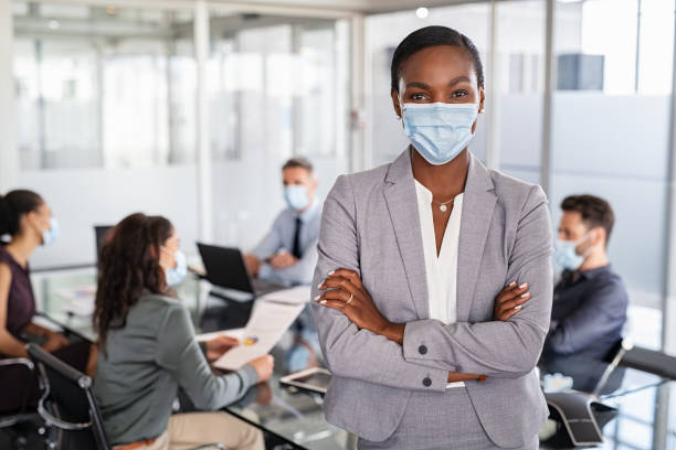 Mature black businesswoman with face mask standing at office Portrait of african american businesswoman wearing protective face mask standing in modern office. Successful black entrepreneur in conference room leaning over table wearing surgical mask with business people working in background. Successful leader looking at camera with crossed arms at work. covid 19 positive stock pictures, royalty-free photos & images