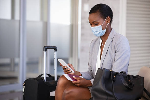 Business woman in airport with face mask checking phone Mature black businesswoman using mobile phone at the airport in the waiting room while wearing face mask due to covid-19. African business woman holding passport and boarding pass typing on smartphone in lounge area during coronavirus pandemic. Female passenger sitting with luggage at terminal hall while waiting for her flight. business travel stock pictures, royalty-free photos & images