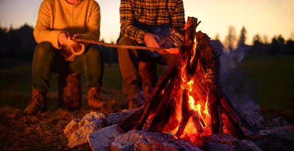 Low section of mature couple roasting sausage over camp fire at dusk.