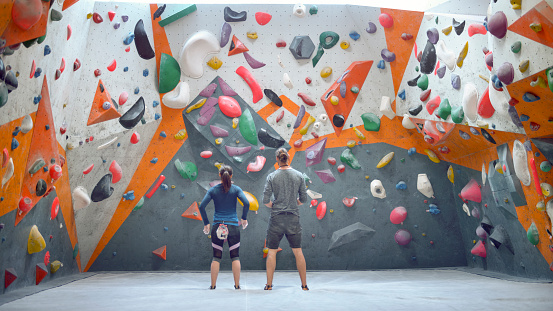 Rear view of instructor and student standing in front of indoor climbing wall in gym.