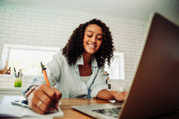 istock Mixed race female with afro studying in kitchen writing on notepad looking at laptop 1308393412