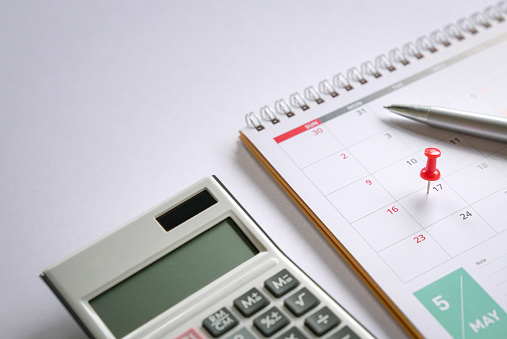 Red push pin on May 17 calendar with pen and calculator on top of table. Reminder of new tax day for 2021.