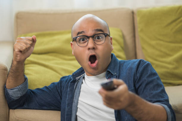 istock Young asian man watching television using remote to change the program. He very enjoy and excited. He's in living room sitting on sofa. 1308394528