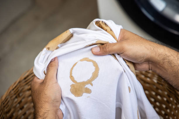 weißes baumwollhemd viele flecken, flecken, kaffeeflecken, die hand des mannes hält das hemd hoch und breitet es aus, um schmutzig auszusehen muss zur waschmaschine gebracht werden - schmutzfleck stock-fotos und bilder