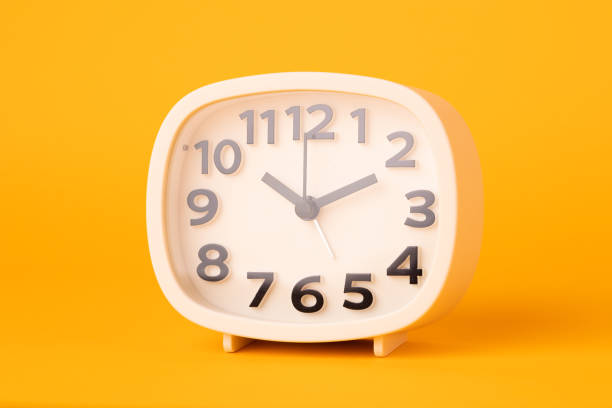 istock Small white alarm clock, black numbers, set the time placed on a table. Clock on isolated yellow background. 1308396453