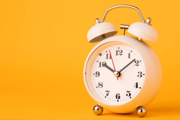 istock Small white alarm clock, black numbers, set the time placed on a table. Clock on isolated yellow background. 1308396465