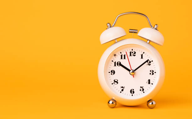 istock Small white alarm clock, black numbers, set the time placed on a table. Clock on isolated yellow background. 1308396468