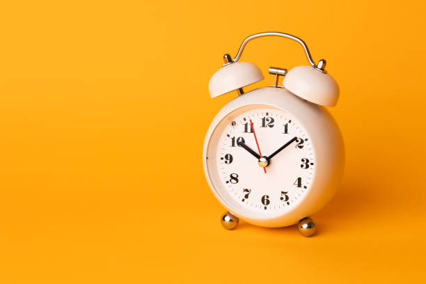 istock Small white alarm clock, black numbers, set the time placed on a table. Clock on isolated yellow background. 1308396470