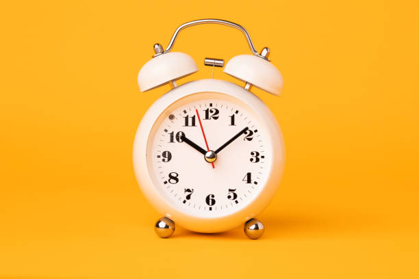 istock Small white alarm clock, black numbers, set the time placed on a table. Clock on isolated yellow background. 1308396482