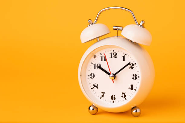 istock Small white alarm clock, black numbers, set the time placed on a table. Clock on isolated yellow background. 1308397240