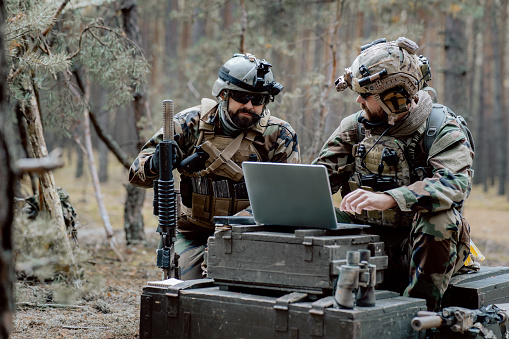 Bearded soldiers in uniform sit on military transport crates, analyze data on a laptop and work out tactics at a temporary forest base. In the background, you can see a soldier protecting the base.