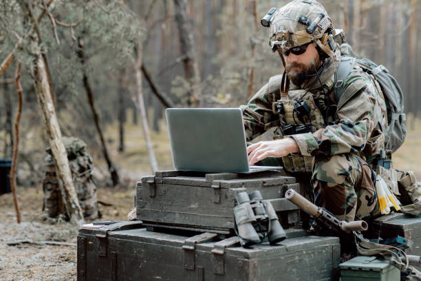 istock Bearded soldier in uniform sit on military transport crates, analyze data on a laptop and work out tactics at a temporary forest base. In the background, you can see a soldier protecting the base 1308397900