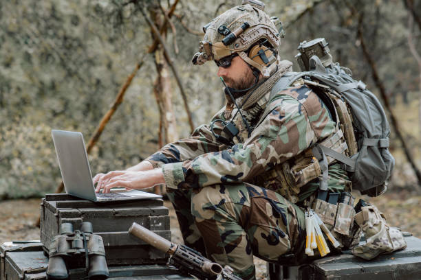 istock Bearded soldier in uniform sit on military transport crates, analyze data on a laptop and work out tactics at a temporary forest base. In the background, you can see a soldier protecting the base 1308398014