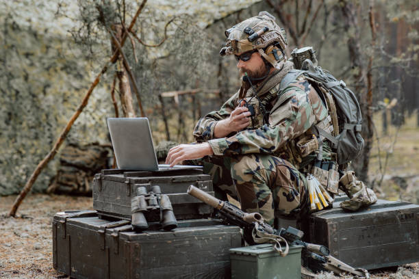 istock Bearded soldier in uniform sit on military transport crates, analyze data on a laptop and work out tactics at a temporary forest base. In the background, you can see a soldier protecting the base 1308398061