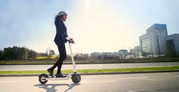 Side view of mature businesswoman riding electric push scooter in city.