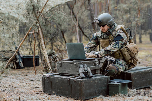 istock Bearded soldier in uniform sit on military transport crates, analyze data on a laptop and work out tactics at a temporary forest base. In the background, you can see a soldier protecting the base 1308398909
