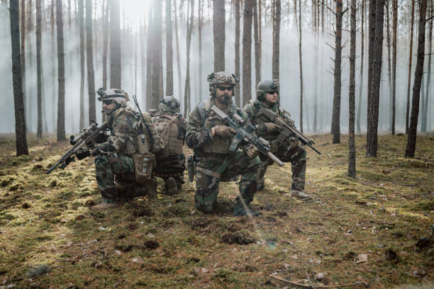 cuatro soldados de mediana edad totalmente equipados con uniformes de camuflaje forman una línea, listos para disparar, apuntando con sus rifles. una operación militar en acción, una unidad de pie en un denso bosque - airsoft fotografías e imágenes de stock