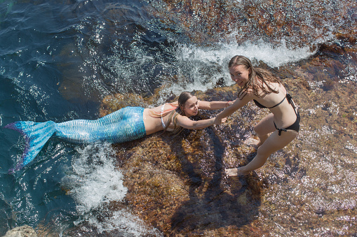 Girl in a black bikini pulls a friend in a mermaid costume out of the water