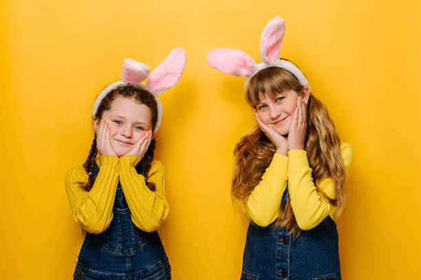 istock Charming lovely little girls keeps hands on cheeks, beautiful sisters with friendly expression, dressed in pink bunny fluffy ears, isolated on yellow studio background. Happy Easter holiday concept 1308400958