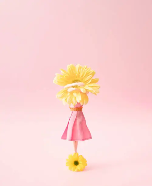 A woman in a pink dress stands on a yellow flower holding a gerbera flower in her hands.