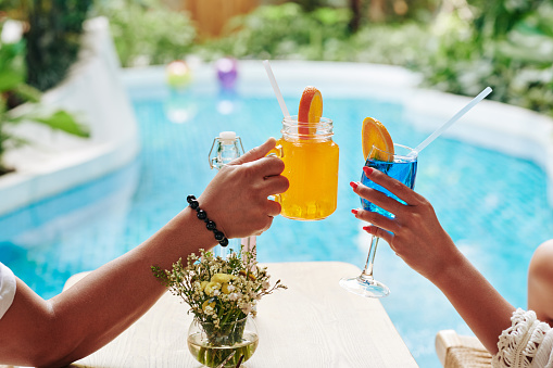 Hands of man and woman clinking glasses of sweet fruit cocktails when enjoying sunbathing by swimming pool