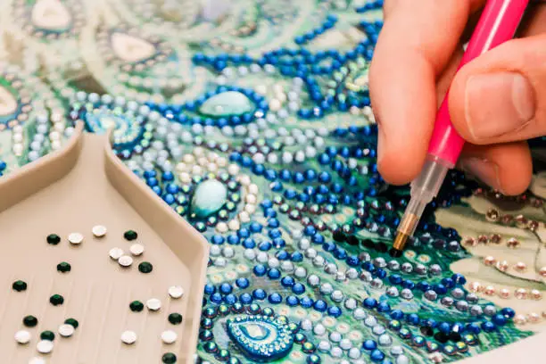 Diamond painting embroidery craft. Acrylic rhinestones and hand holding a pen. Closeup, selective focus.