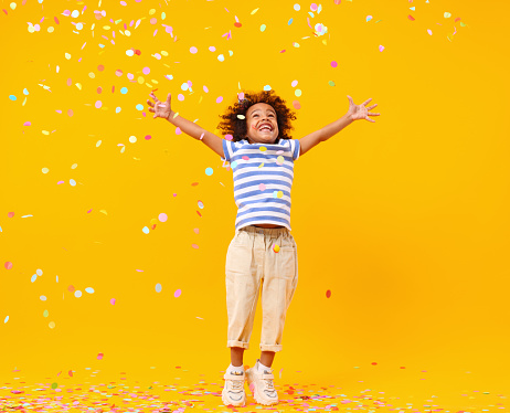 Full body of joyful little black child with Afro hair in stylish clothes laughing and jumping while trying to catch colorful confetti on birthday against yellow background