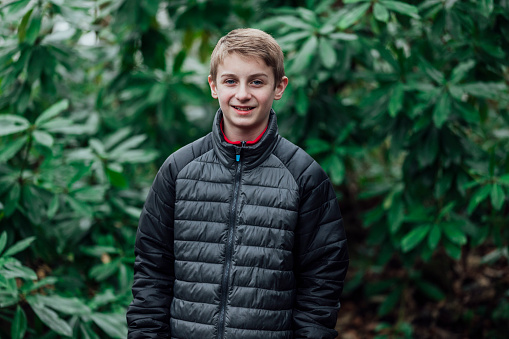 Portrait of a teenage boy standing outdoors in a woodland area while smiling at the camera. He is dressed warmly in winter and is in the North East of England.
