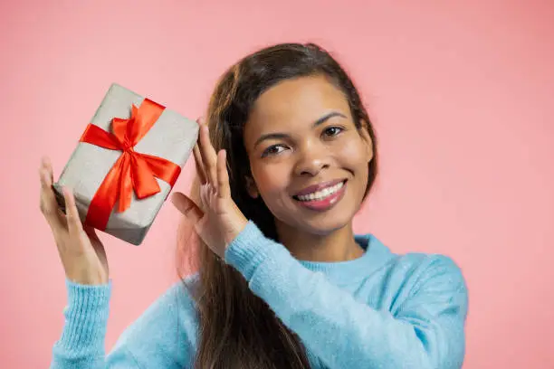 Excited african woman received gift box with bow. She is happy and flattered by attention. Girl smiling with present on pink background. Studio portrait.