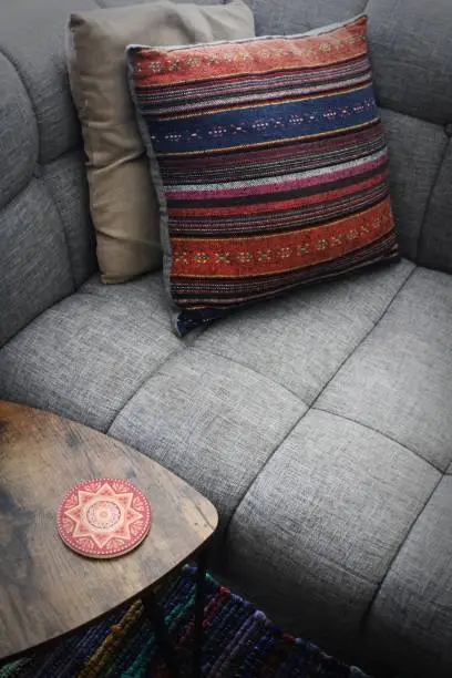 An orange and blue woven throw pillow and coordinated coaster accent the grey upholstery of this sofa and brown end table.