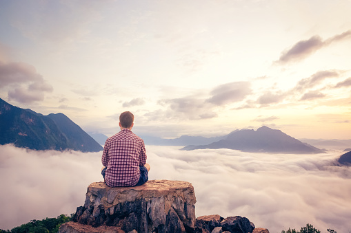 A man sitting on a rock with a beautiful view above a valley filled with clouds. The sun is just below the horizon casting a warm light into the sky.