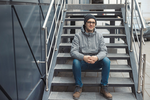 Closeup portrait of handsome business man 30-35 outdoors studying, working, holding a book, planner, wearing gray jacket, hat, jeans, glasses, smiling and sitting on the stairs. Street style photo.