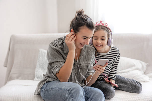 una joven madre y su hija están escuchando música en sus teléfonos mientras están sentados en el sofá de su casa. - mothers audio fotografías e imágenes de stock