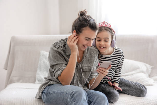 a young mother and her daughter are listening to music on their phones while sitting on the couch at home. - parent mother music listening imagens e fotografias de stock