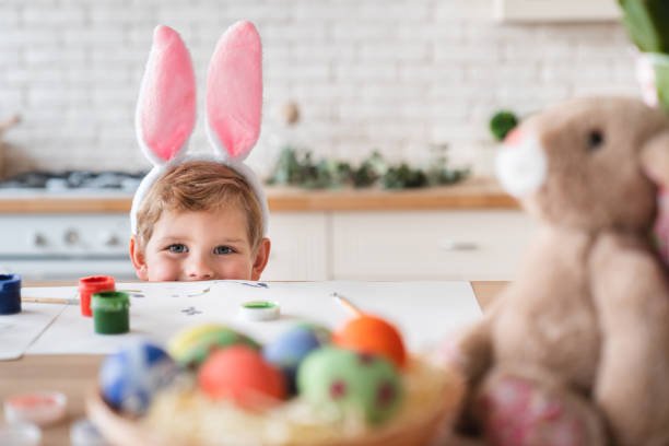 istock Playful little boy in bunny ears hiding behind the table with Easter eggs 1308441770